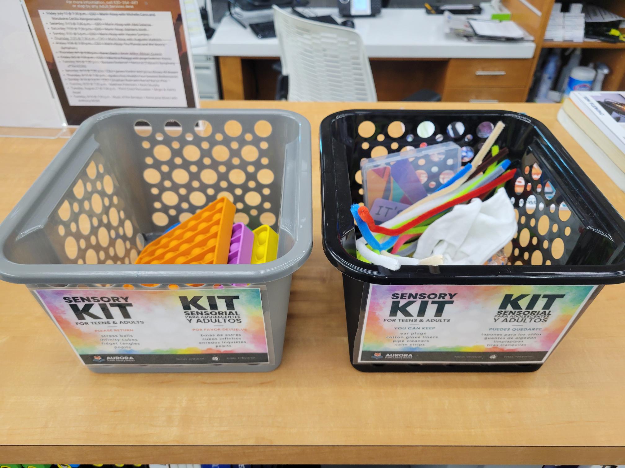 Two bins side by side sit on a table. The gray bin on the left has pop it toys in orange, purple and yellow. The black bin on the right has twist ties of various colors.