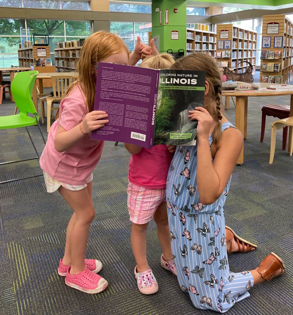 Three children pose with their faces deep in a book at the library