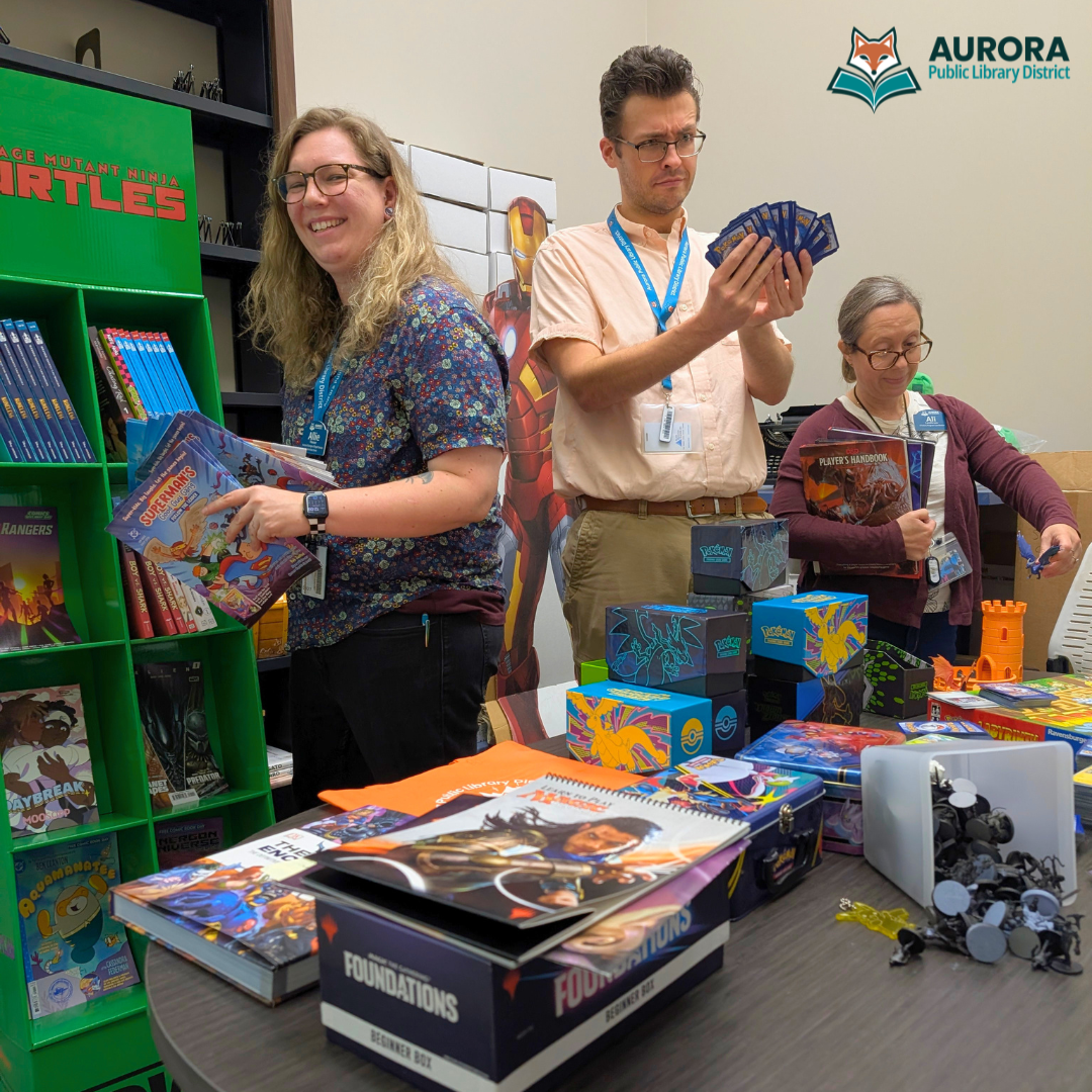 Three staff members prepare comic books, trading cards, toys, and dragons while a cardboard cutout of Iron Man supervises from the corner.