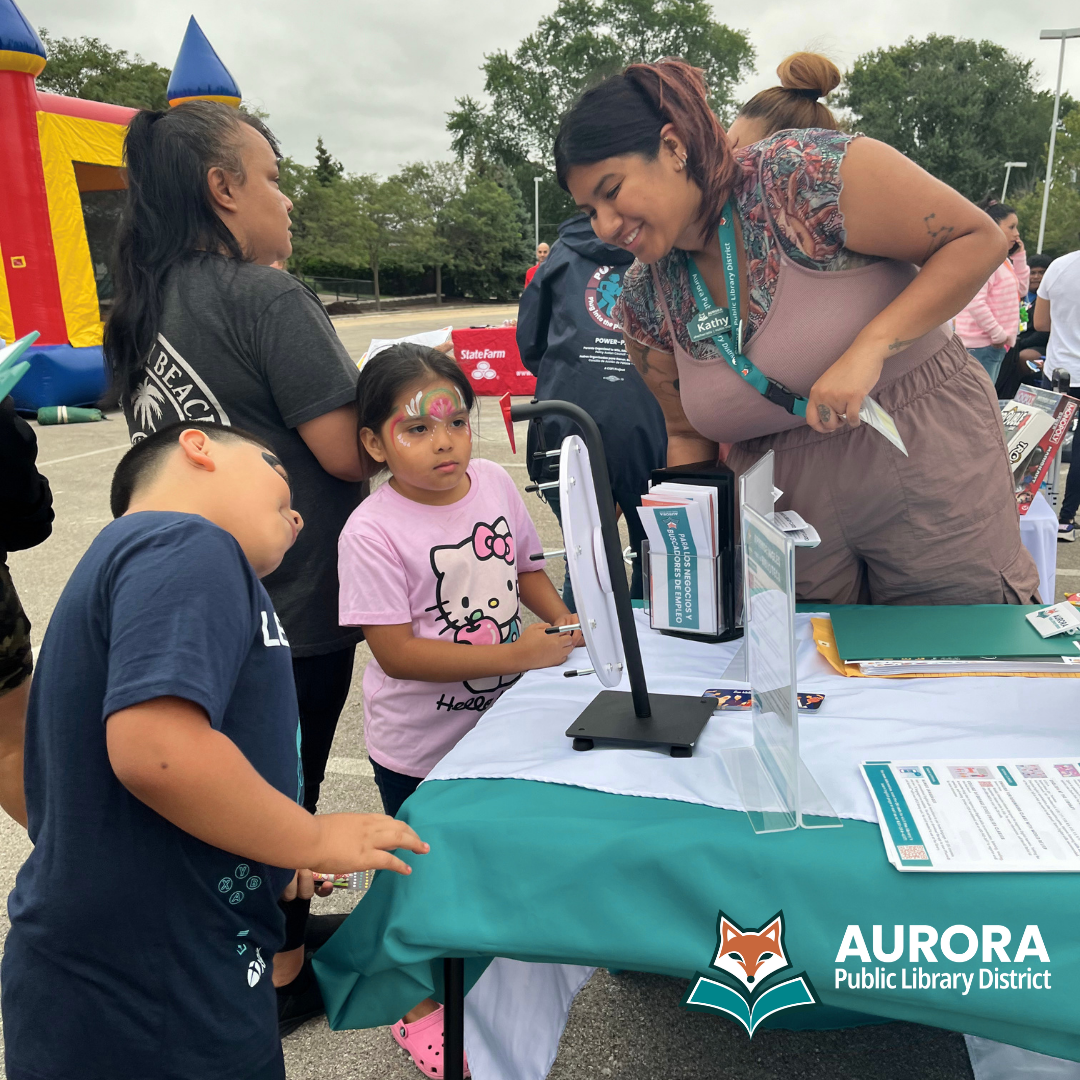 Library staff member at a table during an outdoor event interacting with young customers who have their faces painted. A bouncy house and a crowd of attendees are in the background.