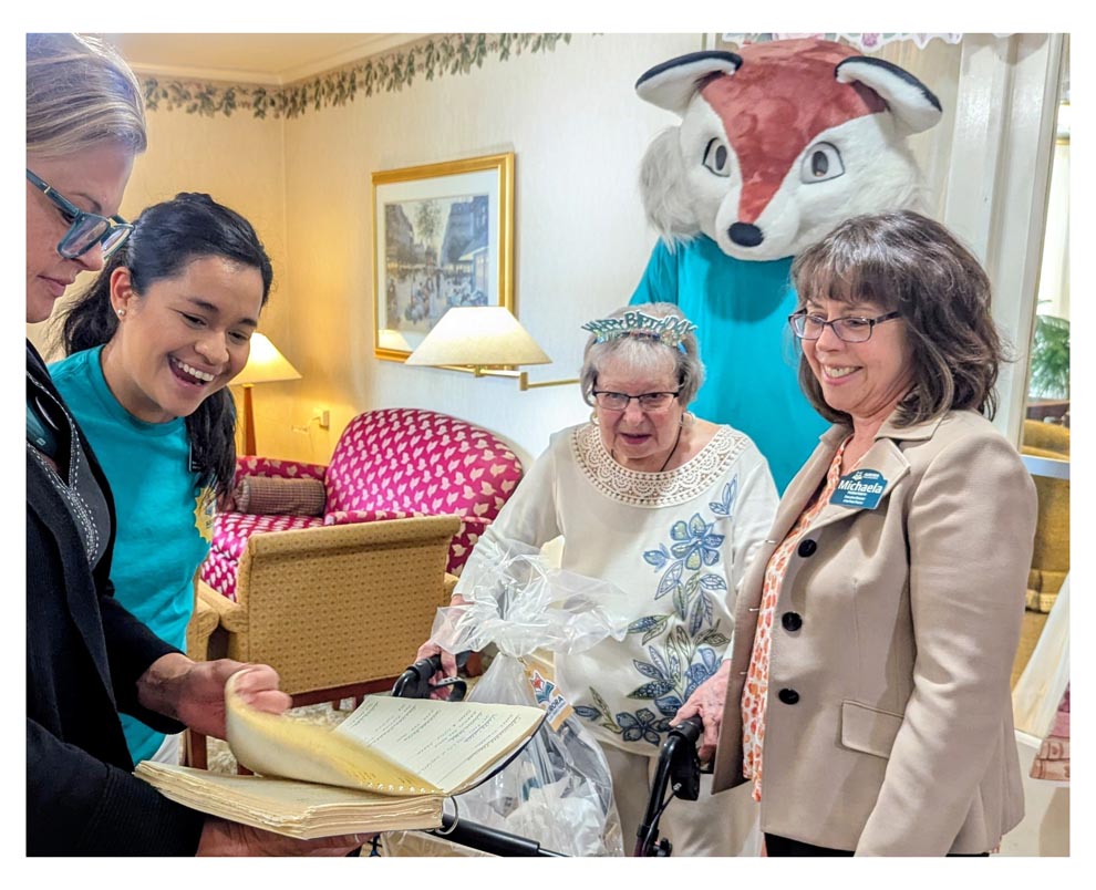 Plum landing staff, library staff, Rory the fox mascot, and Jennie wearing a birthday headband gather to look at a journal.