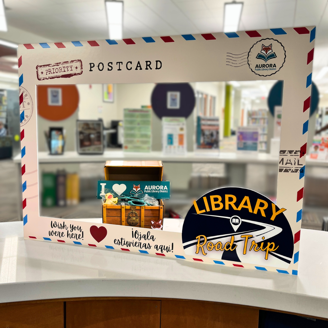 A large postcard themed photo frame with the Library Road Trip logo sits next to a treasure chest of APLD branded giveaways.