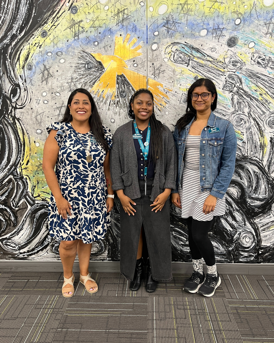 Staff members stand and pose and smile in front of the detailed mural at the Santori Library.