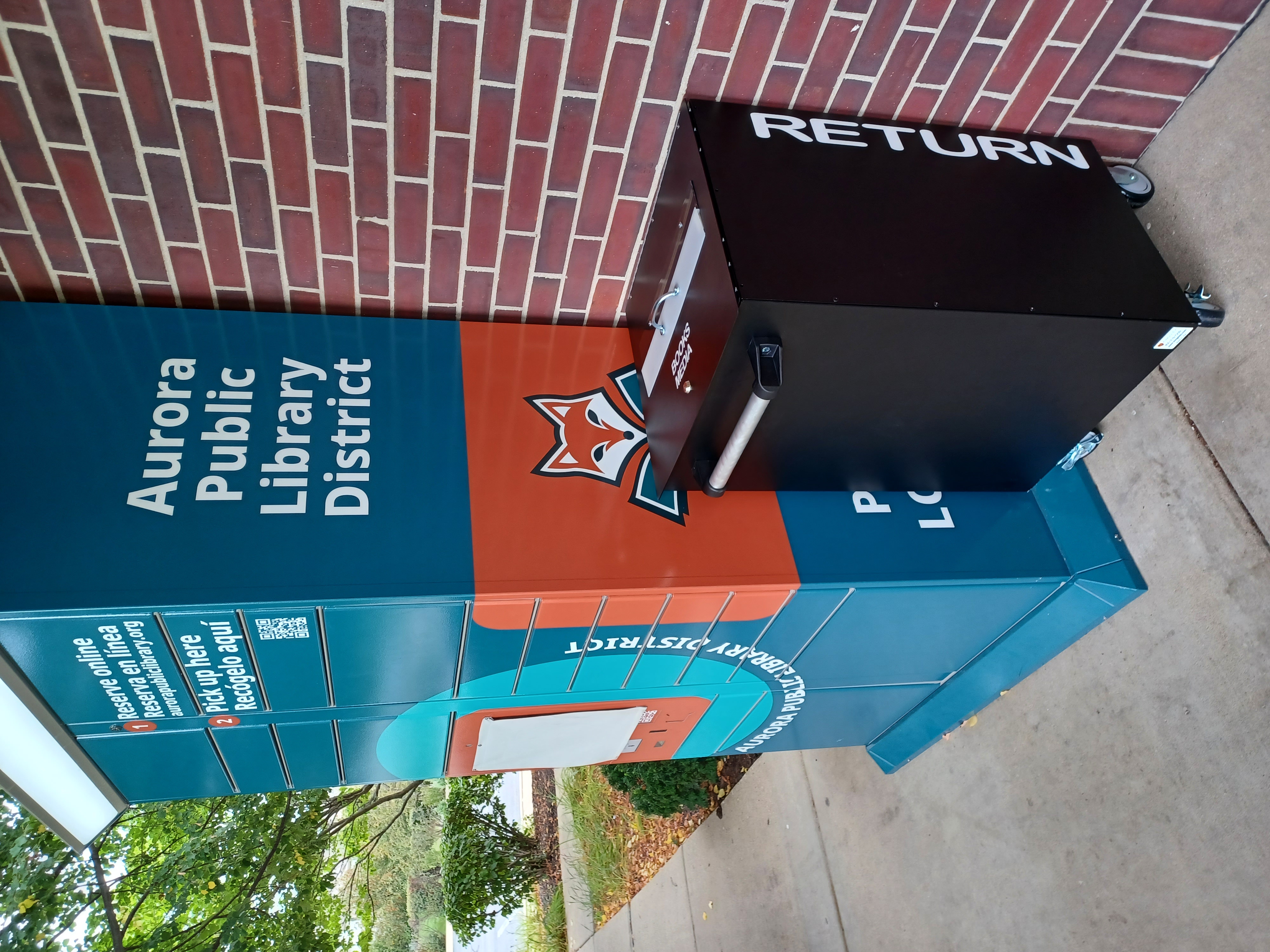 Teal and orange outdoor APLD pickup locker with a mobile return cart next to it.