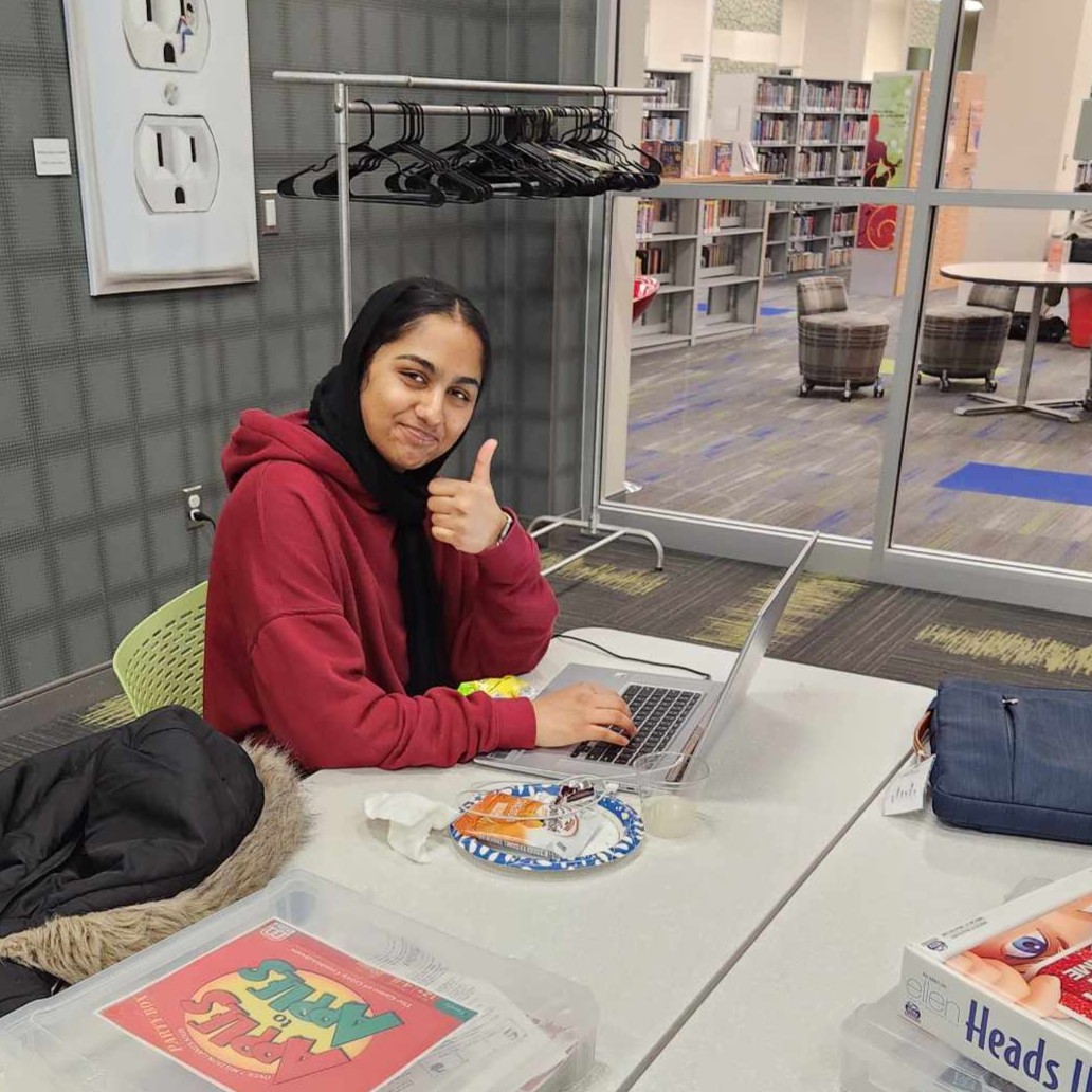 Teen gives a thumbs up to the camera while working on a laptop during a TAB event at the library..