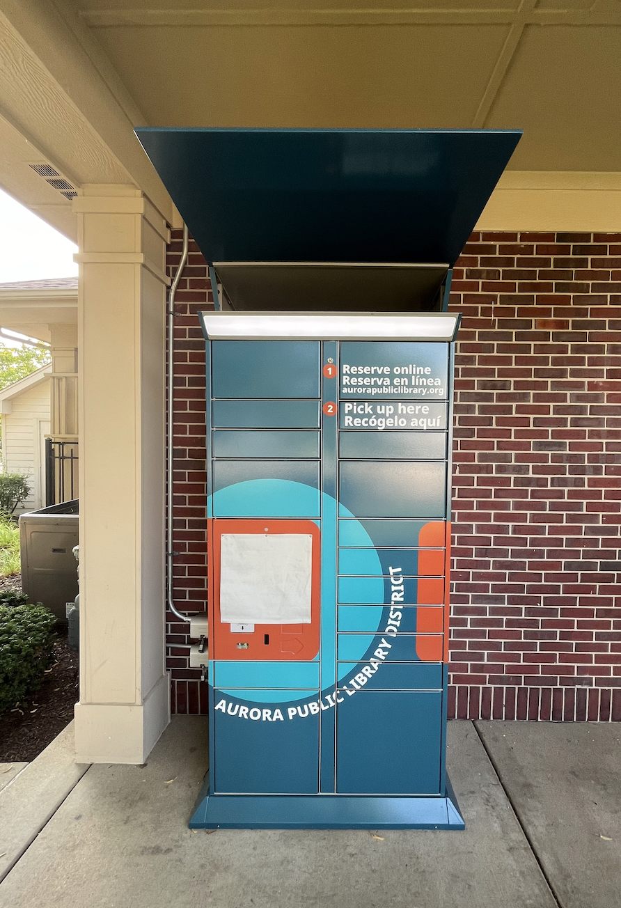 Orange and teal pickup APLD pickup locker outdoors against a brick building.